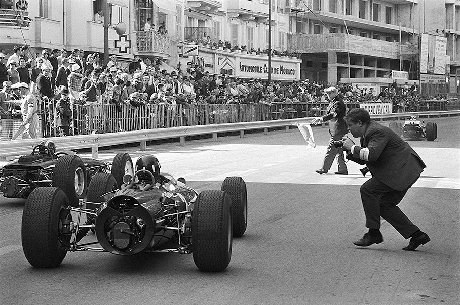 Pole position, fastest lap and third victory in Monaco 1965 for Graham Hill, here on his honoury lap. Former Grand Prix driver Louis Chiron, now race director, waves a flag and still lives a dangerous life - like the photographer Horst Baumann.