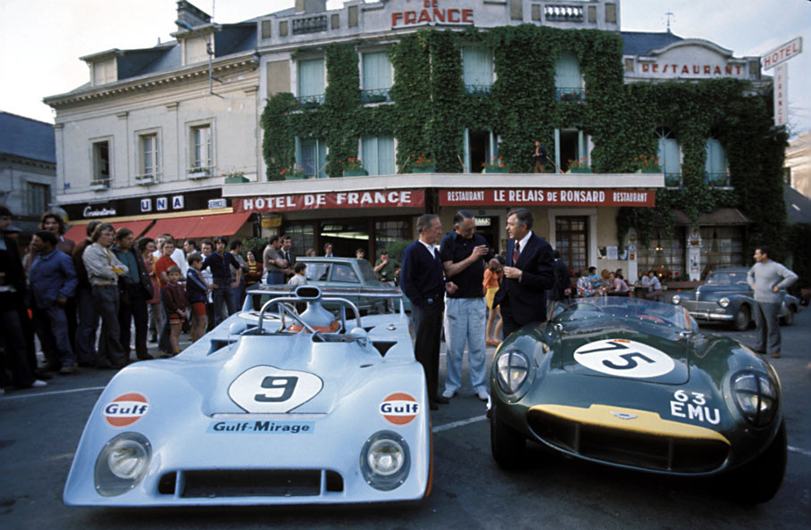 John Wyer next to an unknown gentleman besides the Gulf Mirage Ford and an Aston Martin DB3 racecar with its owner Richard Pilkington in front of Hôtel de France in Chateau du Loir