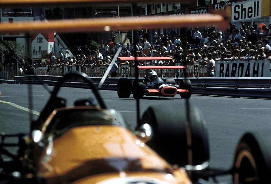 John Surtees, BRM, seen through the huge wings of a McLaren, passes start/finish. After the first practice the CSI ban on the grounds of safety the high rear wings used on the cars.