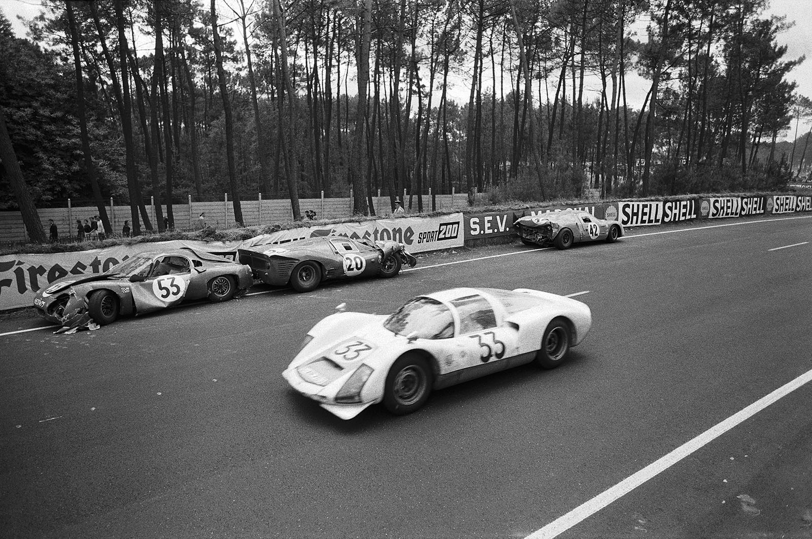 A Porsche 906 155 S-XR 763, driven by Peter Gregg (USA) and Sten Axelsson (S), overtakes the wrecks of three retired racecars besides the track. Car no. 42 is the Matra M620 BRM 03 driven by Jo Schlesser (F) and Alan Rees (GB), car no. 20 the Ferrari 330 P3 #0848 of Mike Parkes (GB) and Ludovico Scarfiotti (I), car no. 53, the C.D. SP66 Peugeot 663IW75 of Georges Heligouin (F) and Johnny Rives (F)